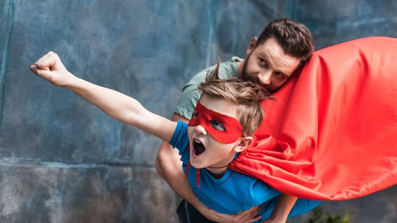 father holding son in superhero costume flying at home